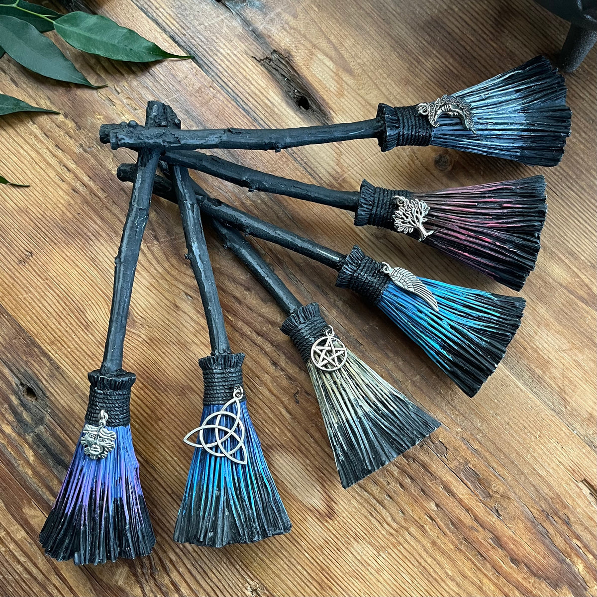 Six miniature witch brooms with dark wooden handles and colourful brush in shades of blue, purple, and pink, arranged on a rustic wooden surface.
