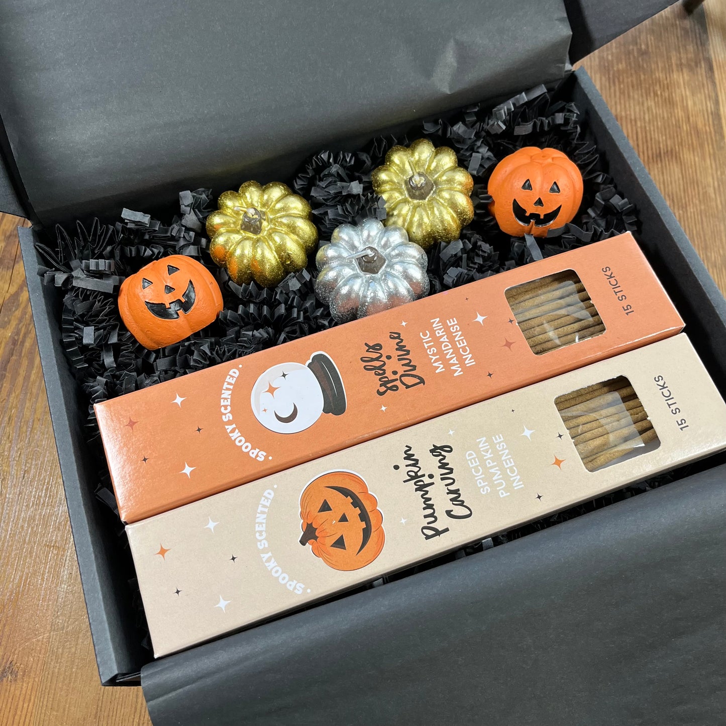 A Halloween-themed gift box containing two boxes of scented incense and three small decorative pumpkin candles and two pumpkin incense holders arranged in black crinkle paper inside a dark gray box.