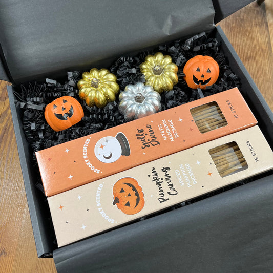 A Halloween-themed gift box containing two boxes of scented incense and three small decorative pumpkin candles and two pumpkin incense holders arranged in black crinkle paper inside a dark gray box.