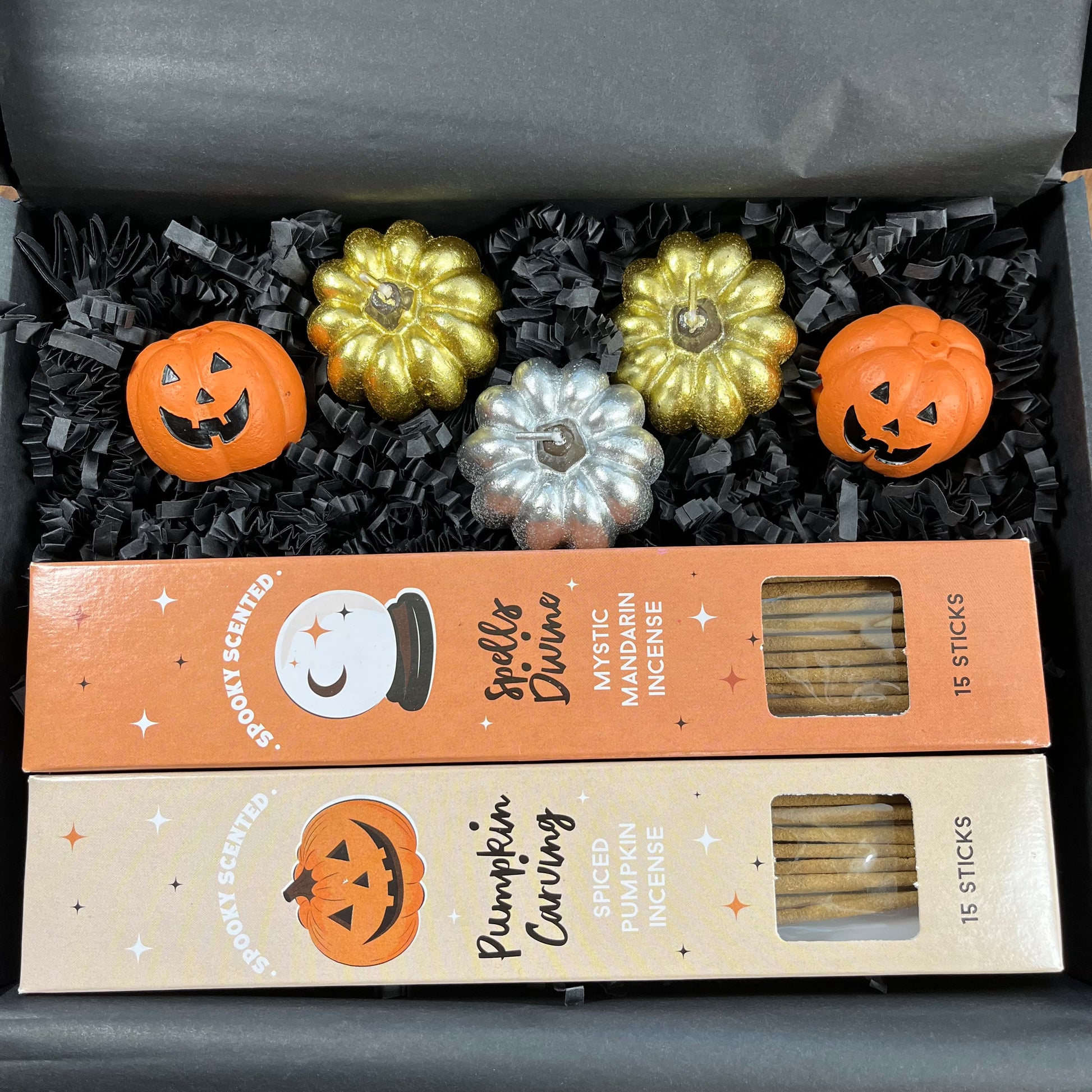 A Halloween-themed incense gift set in a black box filled with black shredded paper. The set includes two boxes of incense sticks labeled 'Spells of Mymics' and 'Pumpkin Carving', along with four decorative pumpkins in orange, gold, and silver.