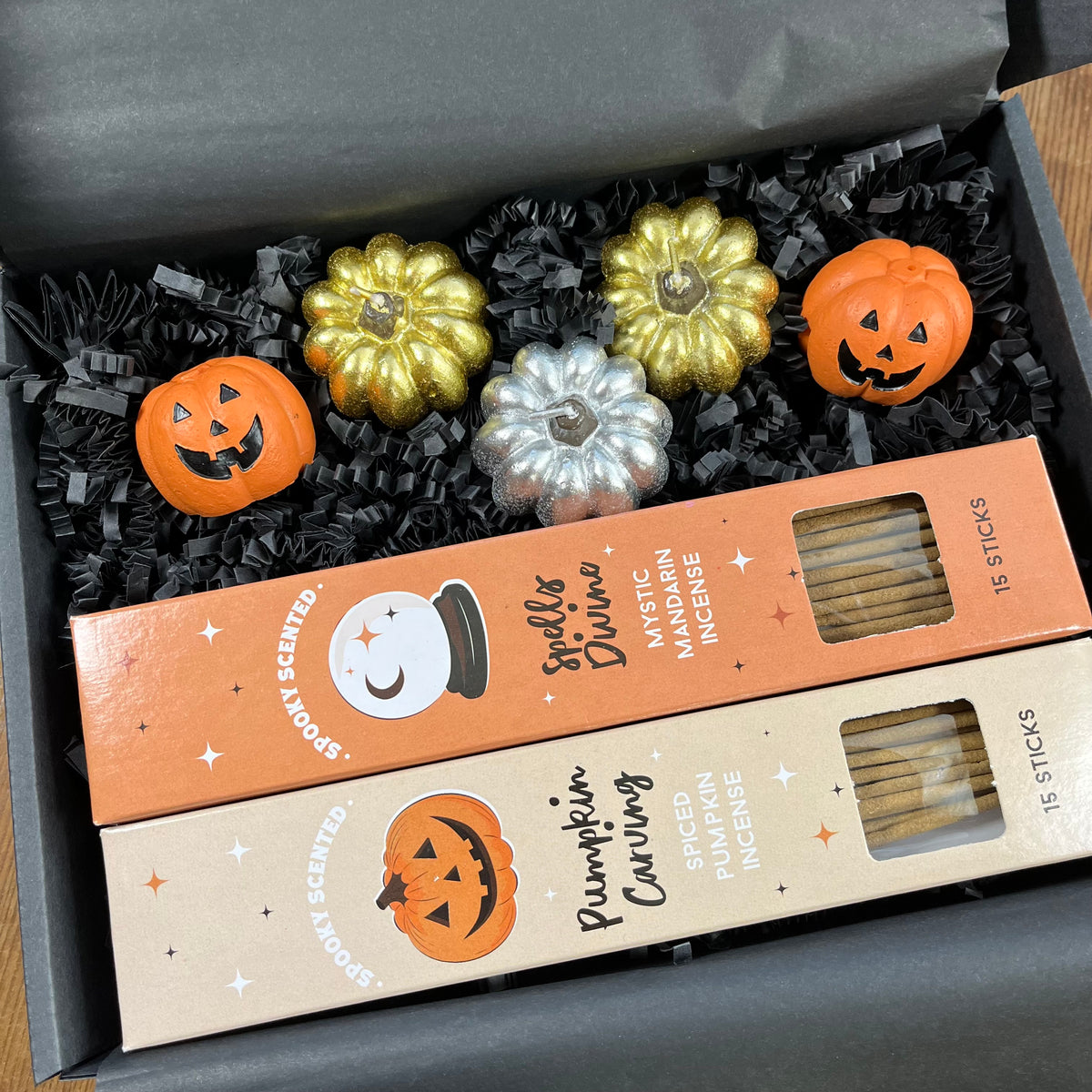 A Halloween-themed scented incense gift set in a dark gray box, featuring two boxes of incense sticks and three decorative pumpkins in gold, silver, and orange pumpkin incense holders resting on black crinkle paper.
