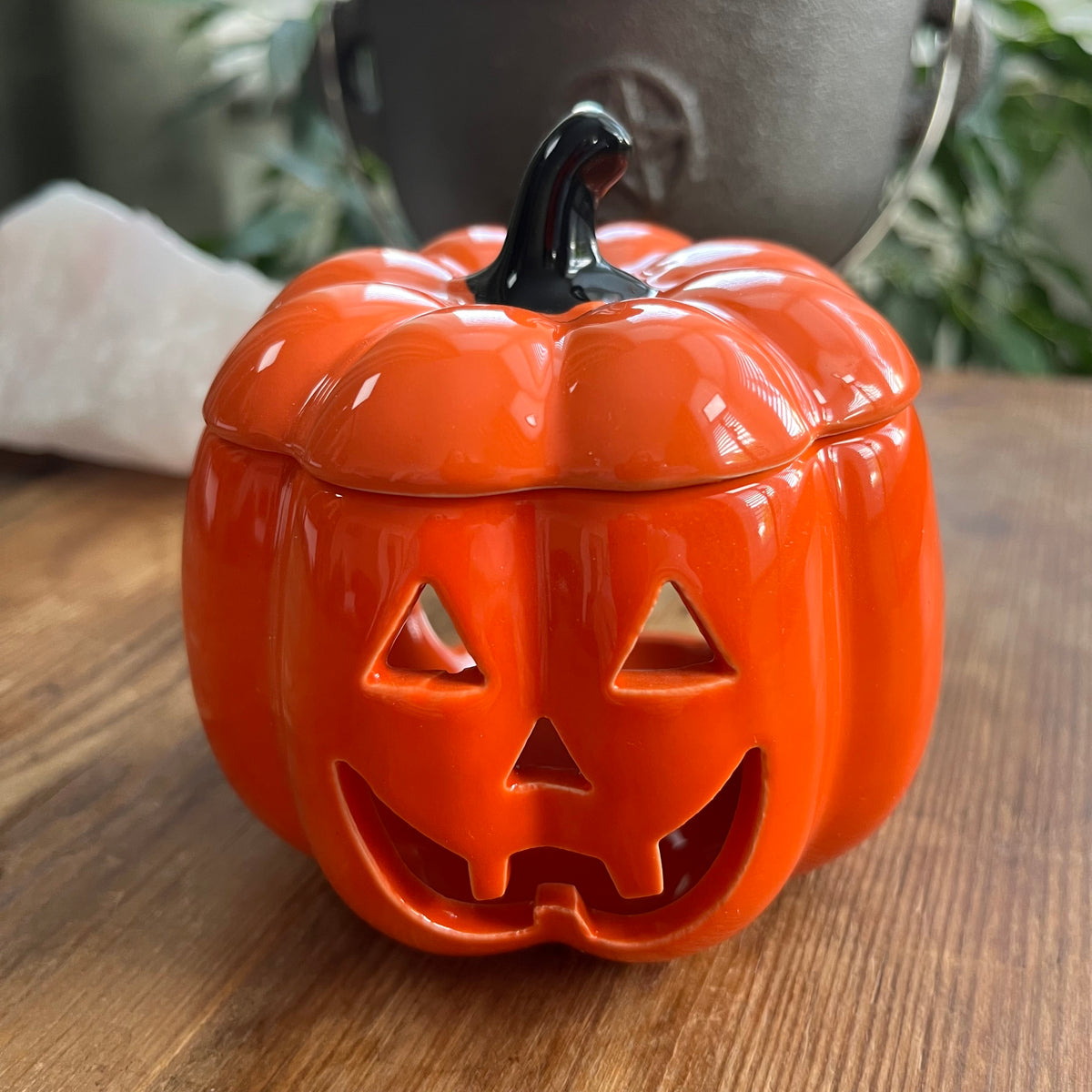 An orange pumpkin-shaped ceramic oil burner with a carved jack-o'-lantern face and a black stem, sitting on a wooden surface.