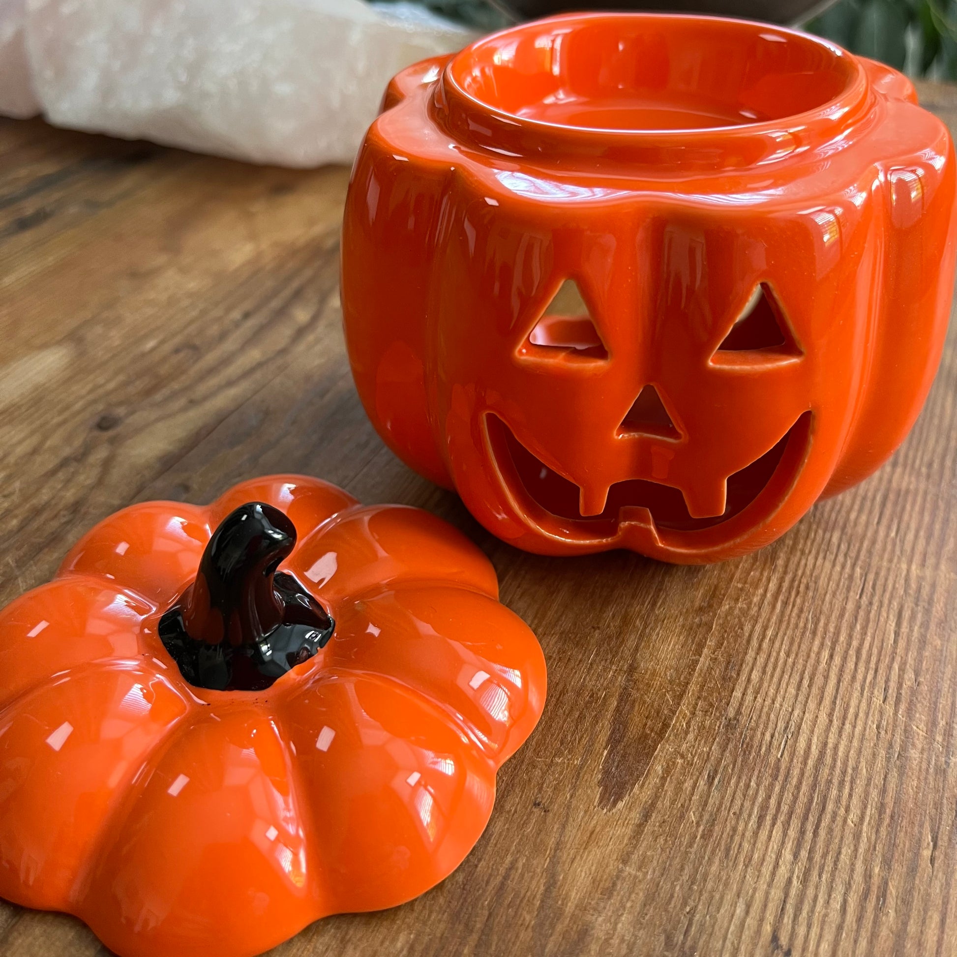 An orange ceramic jack-o'-lantern oil burner with its lid placed in front on a wooden tabletop. The glossy pumpkin-shaped container has carved facial cutouts 