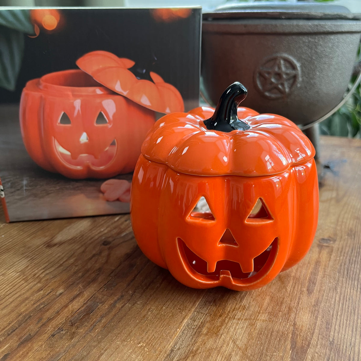 An orange pumpkin-shaped ceramic oil burner with a carved jack-o'-lantern face and a black stem, shown on a wooden table with its product box in the background.