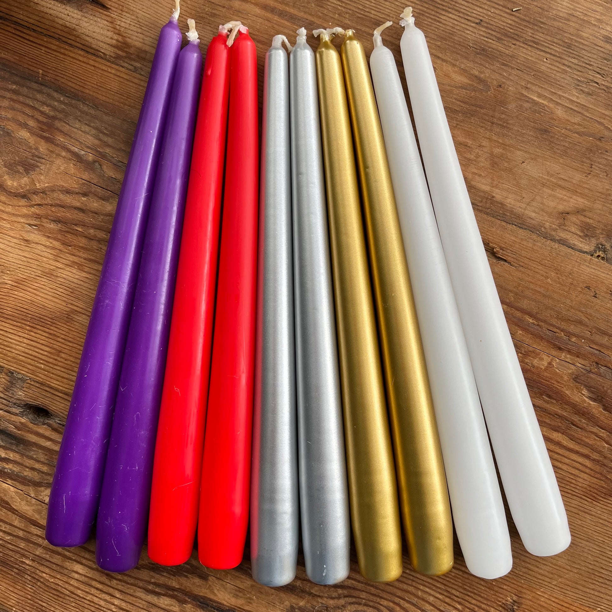 A set of eight taper candles in assorted colors, including purple, red, silver, gold, and white, arranged in a fan shape on a wooden surface.