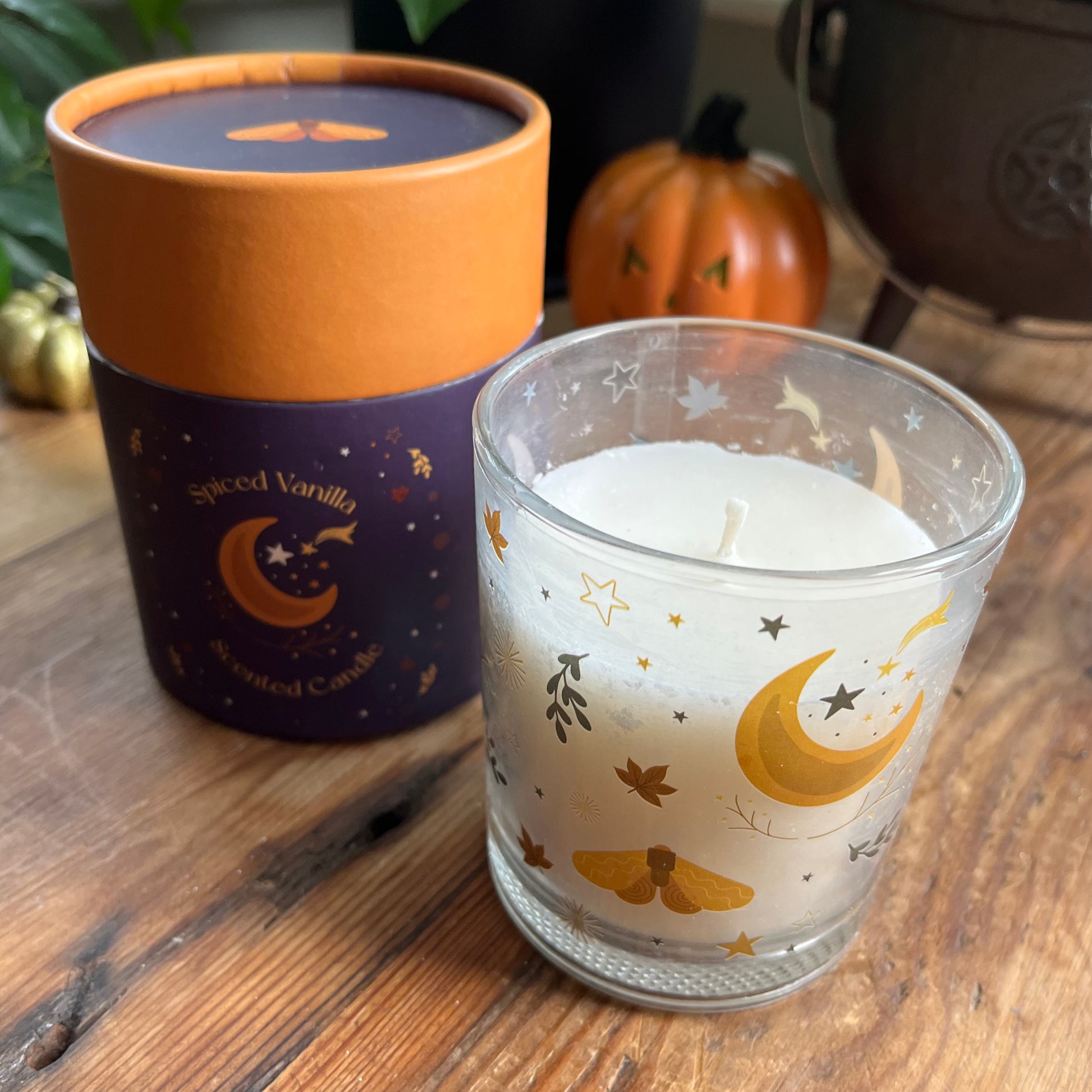 Candle in a glass holder with decorative design on a wooden surface, next to a cylindrical container labeled 'Spiced Vanilla'.