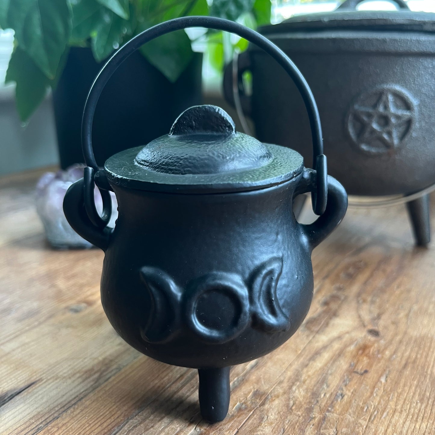 Black cast iron cauldron with lid and triple moon design on a wooden surface, with another cauldron in the background featuring a pentacle design.