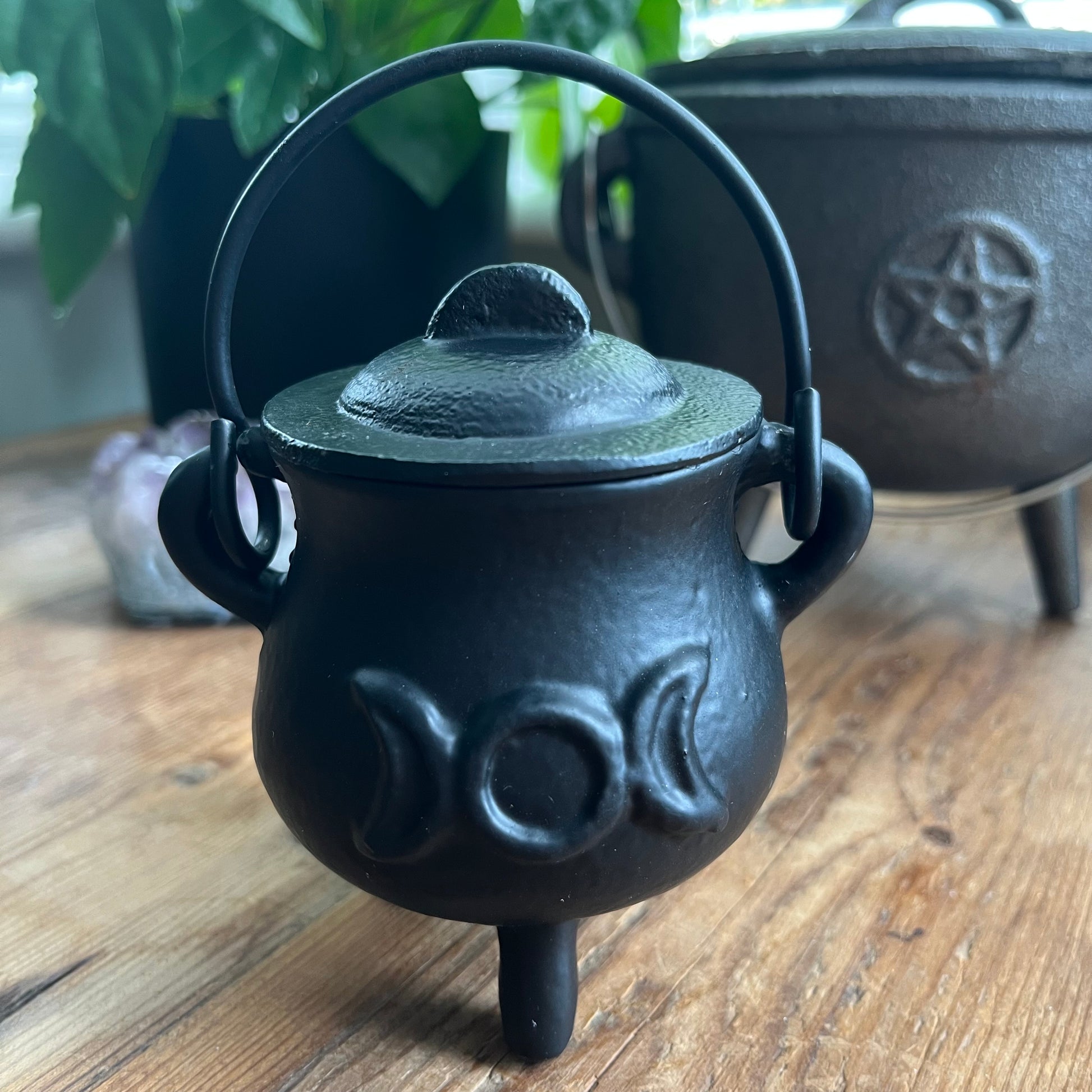 Black cast iron cauldron with lid and triple moon design on a wooden surface, with another cauldron in the background featuring a pentacle design.