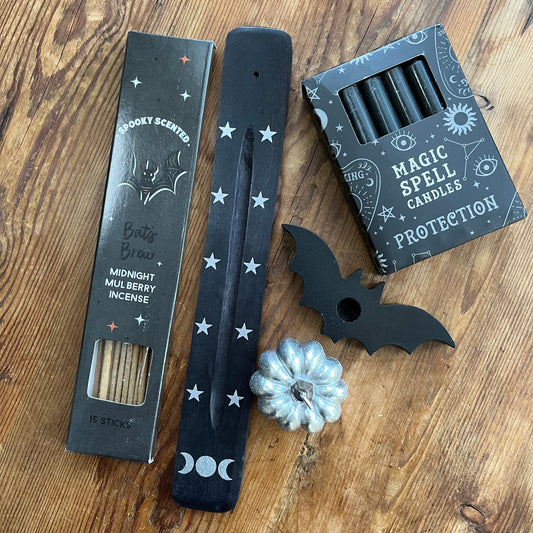 A flat-lay of Halloween-themed witchcraft accessories on a wooden surface, including a box of 'spookey Scented' Bat's Brew Midnight Mulberry Incense sticks, a black wooden incense holder with silver stars, a black 'Magic Spell Candles Protection' box, a black bat-shaped spell candle holder and a small silver pumpkin charm.