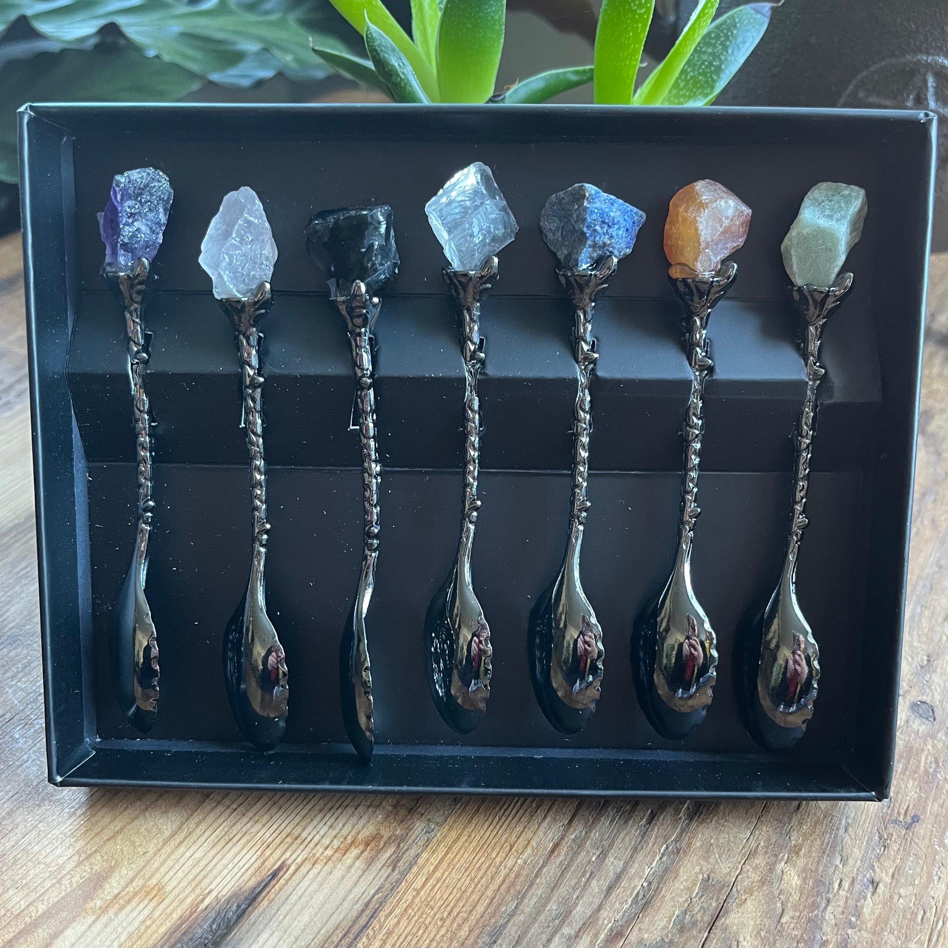 Set of eight decorative spoons with embedded stones in a black box on a wooden surface.