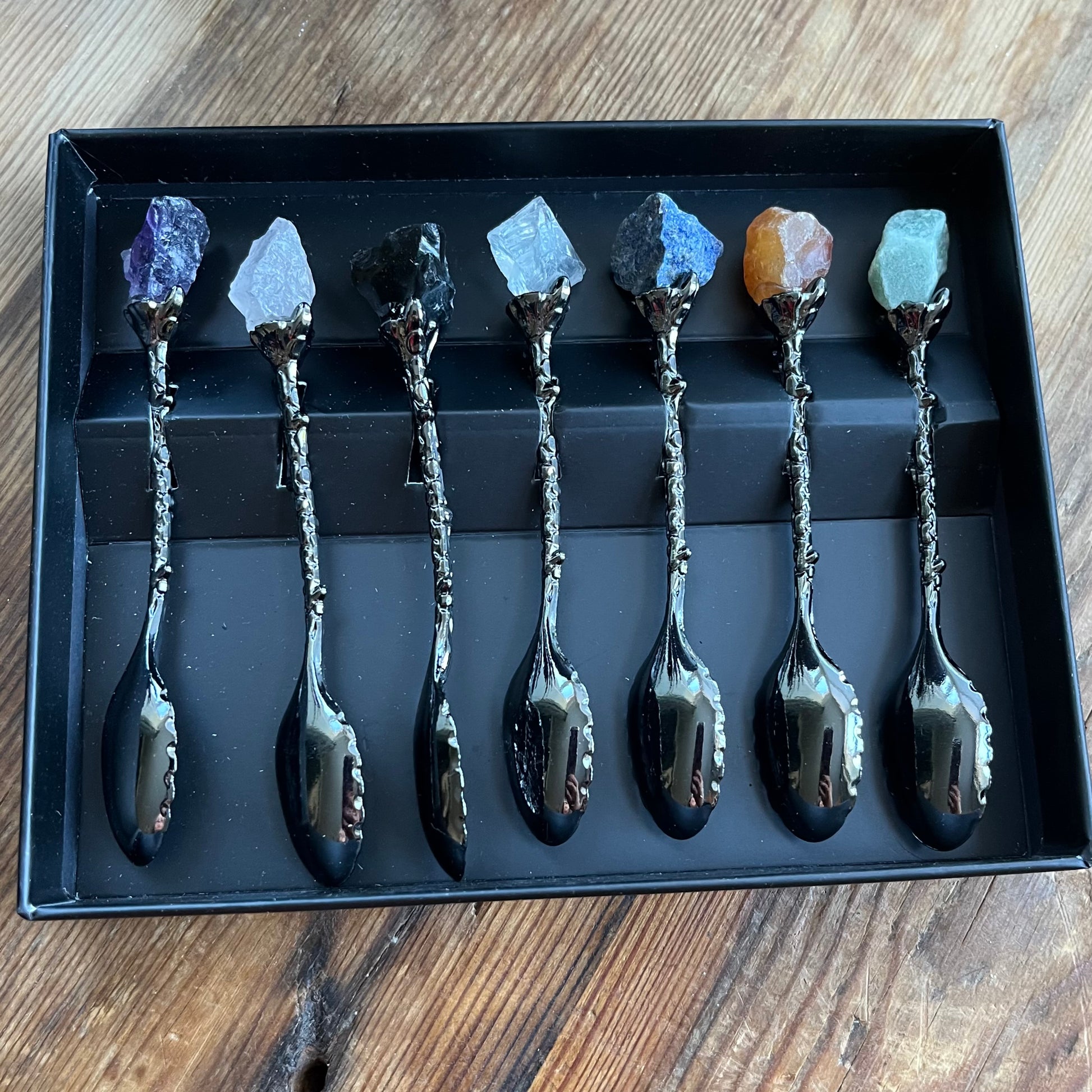 Set of eight silver spoons with gemstone handles in a black box on a wooden surface